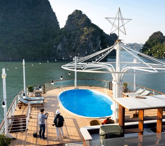 A cruise ship deck with a small oval pool, lounge chairs, and two people looking out at mountainous islands.
