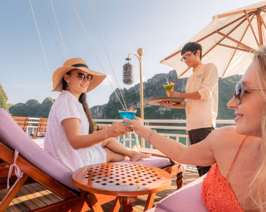 Two women toasting with blue and green cocktails on a sun deck overlooking the scenic islands of Halong Bay.