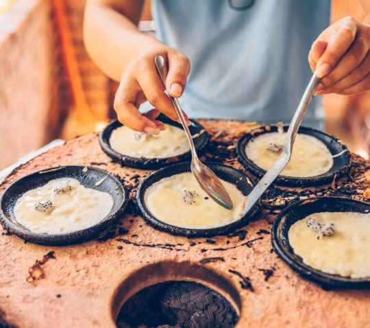 Close-up of hands using spoons to cook small rice batter pancakes in individual pans over hot coals.