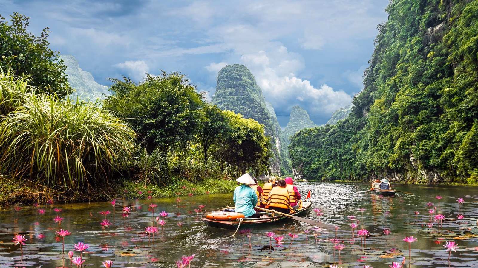 Several small boats rowing through a pond filled with pink lilies, surrounded by large green mountains.
