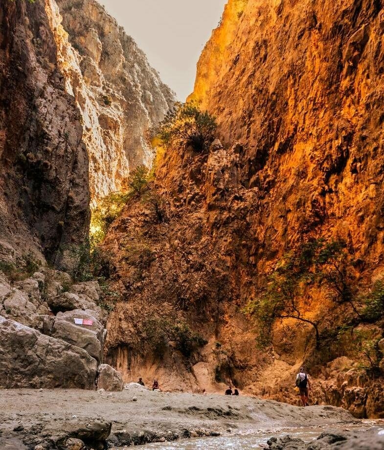 People hiking through a deep, narrow canyon with massive orange rock faces in Samaria Gorge National Park.