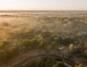 Aerial view of a safari lodge nestled in a lush forest covered in golden morning mist by a winding river.