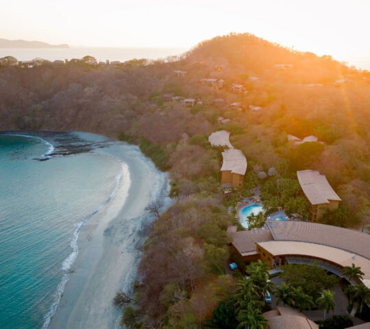 Aerial view of a tropical peninsula and resort buildings during a bright orange sunset.
