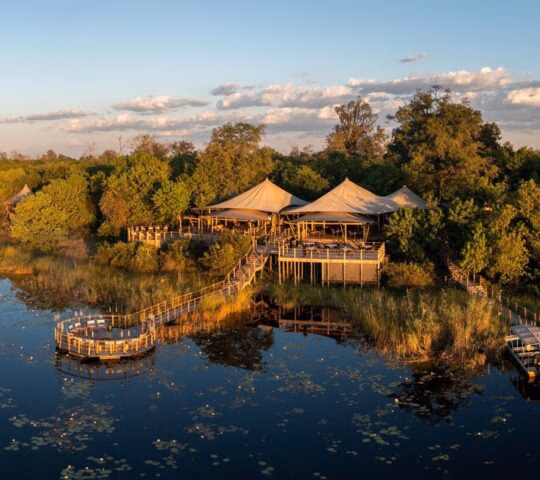 Aerial view of Wilderness DumaTau safari camp with canvas tents and a circular wooden deck over a lily-covered lagoon.