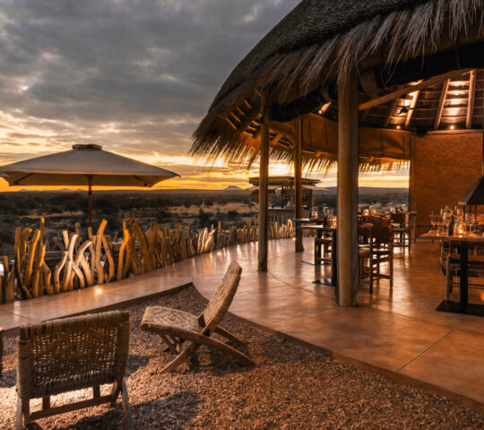 Luxury open-air restaurant at Zannier Hotels Omaanda with thatched roof and tables overlooking a sunset savanna.