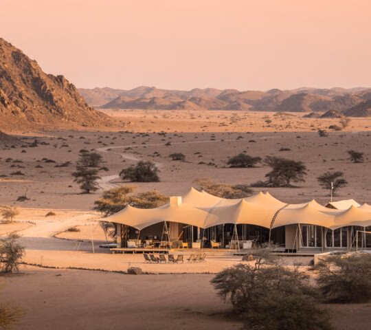 Tented roofs of the luxury Hoanib Skeleton Coast Wilderness Camp in a vast desert valley surrounded by rocky hills.