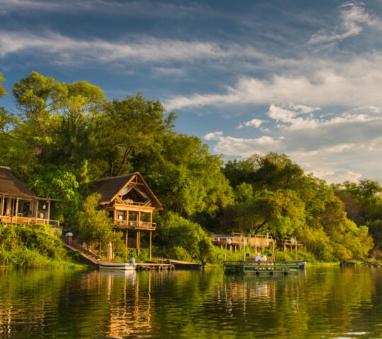 Thatched riverfront lodges at Tongabezi in Zambia surrounded by green trees and reflected in the Zambezi River.