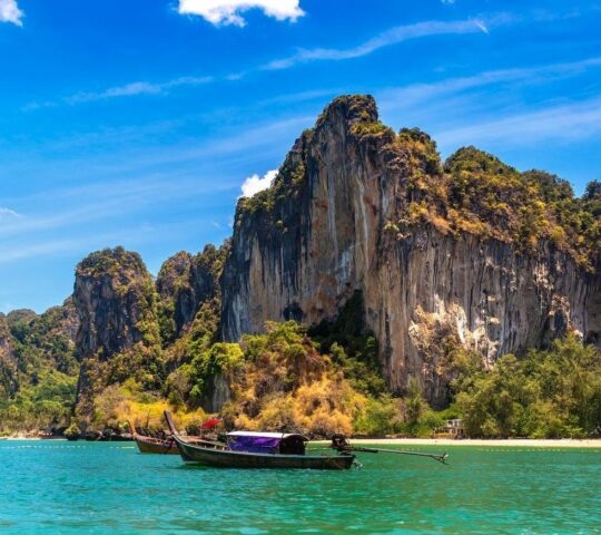 A long boat sailing past a steep limestone cliff at Railay beach in Thailand