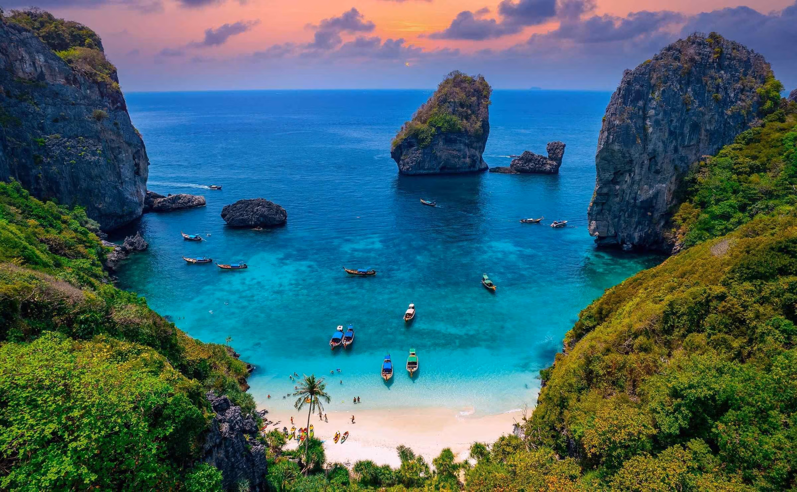 aerial view looking past tree covered cliffs onto a beach with turquoise sea and sunset sky