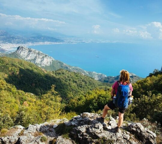 A woman wearing a backpack stop on top of a ridge admiring a view over the Amalfi Coast