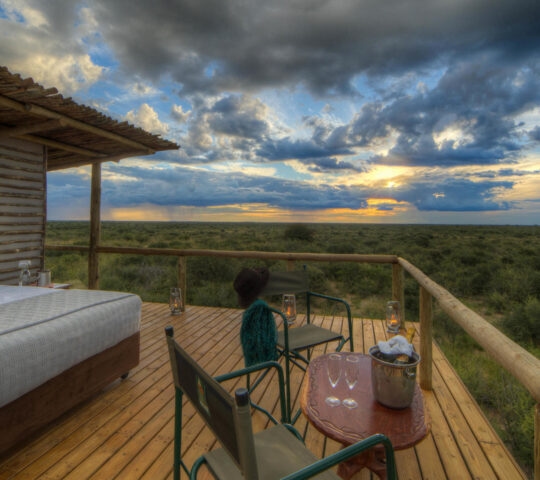 A bed and seating area on a wooden deck at Dinaka Botswana property overlooking a sunset over the Kalahari bush.