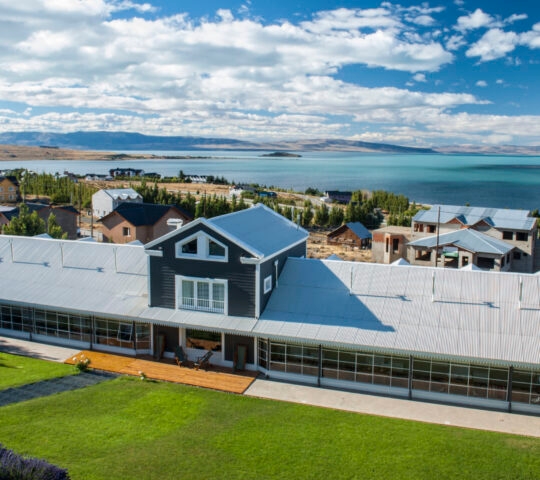 Aerial shot of the hotel with a green lawn in the foreground and scattered houses and trees in the background and the lake and shoreline beyond.