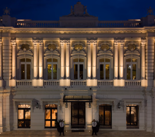 Exterior night view of the hotel with grand colonnades uplit by soft lighting.