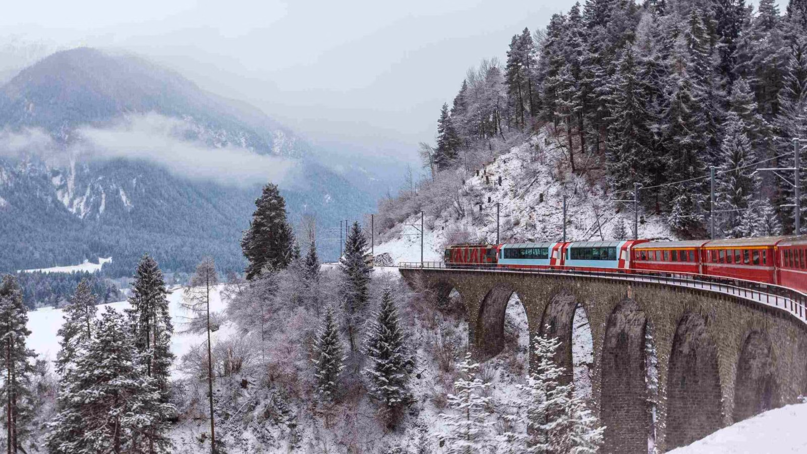 A red passenger train crosses a tall stone viaduct bridge in a snowy mountain forest in Switzerland.