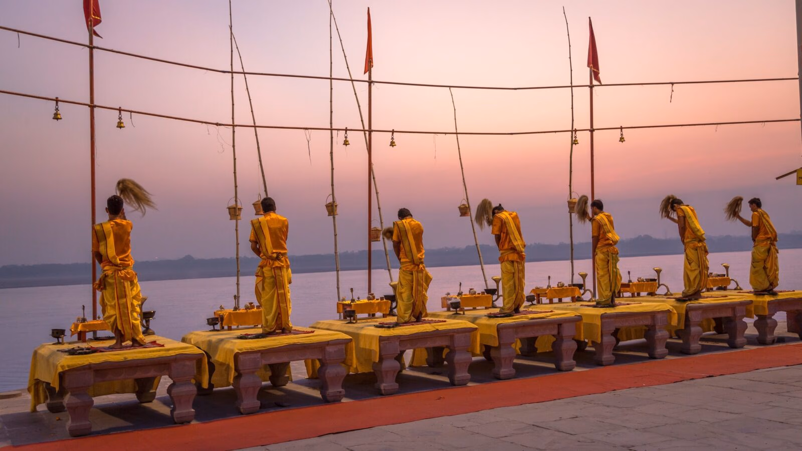 Priests in yellow robes perform a ritual with whisks on platforms overlooking a wide river during a pink sunset.