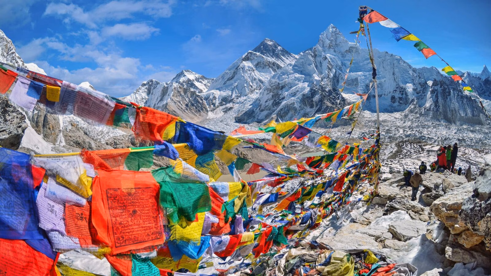 Colorful prayer flags stretch across a rocky mountain pass with massive snow-covered Himalayan peaks in the background.