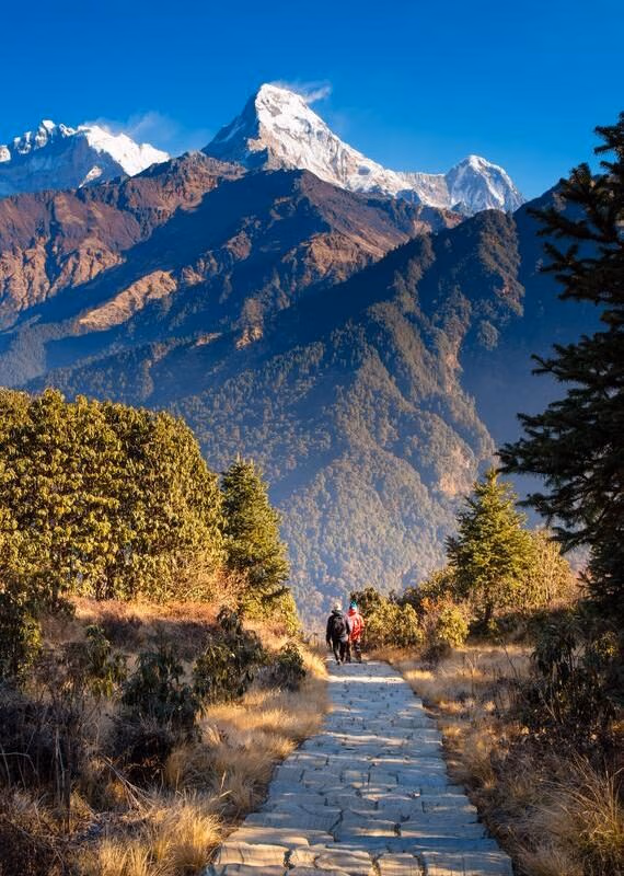 Two trekkers walk on a stone trail toward a large snow-covered mountain peak under a clear blue sky.