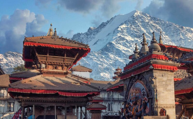 Ornate wooden temples with red trim in a city square with a massive snow-capped mountain rising in the background.