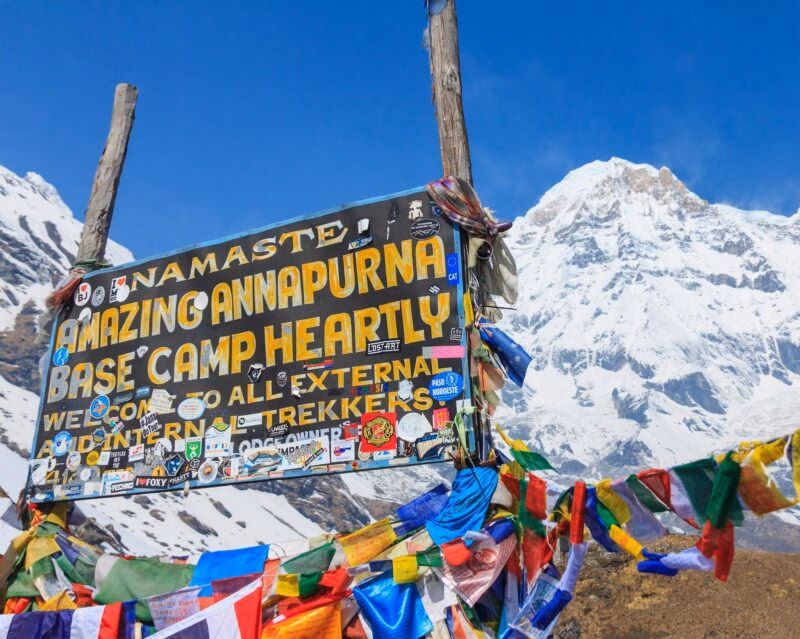 A sign reads Namaste Amazing Annapurna Base Camp Heartly Welcome, covered in stickers with snowy mountains behind it.
