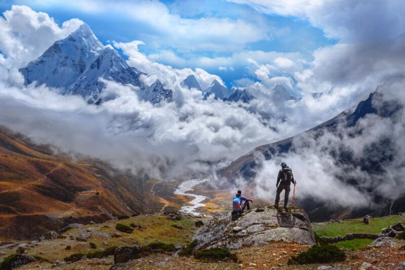 Two trekkers stand on a rock overlooking a deep valley with snow-capped mountains peaking through thick white clouds.