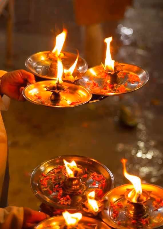 Multiple burning oil lamps on metal trays decorated with red flower petals held during a religious Aarti ceremony.