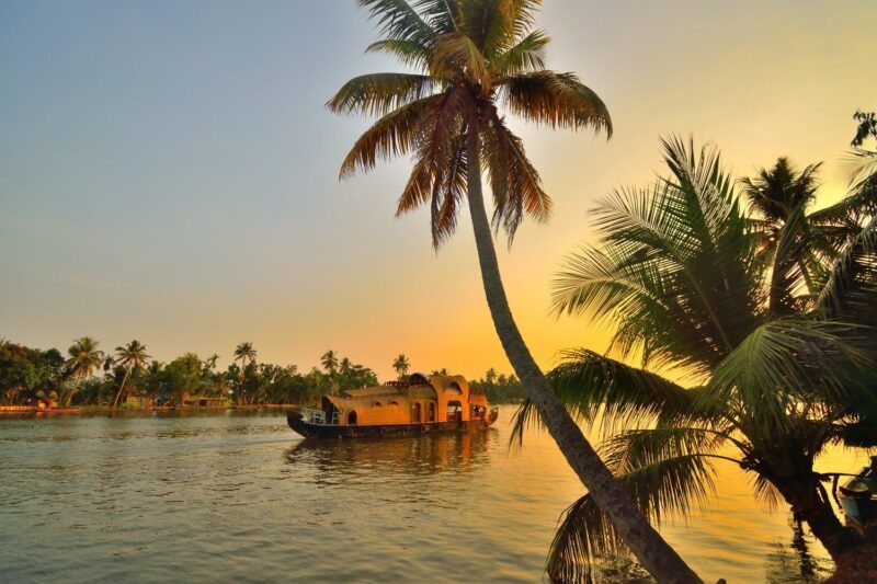 A traditional Kerala houseboat sails on a river at sunset, framed by silhouetted palm trees under a golden sky.