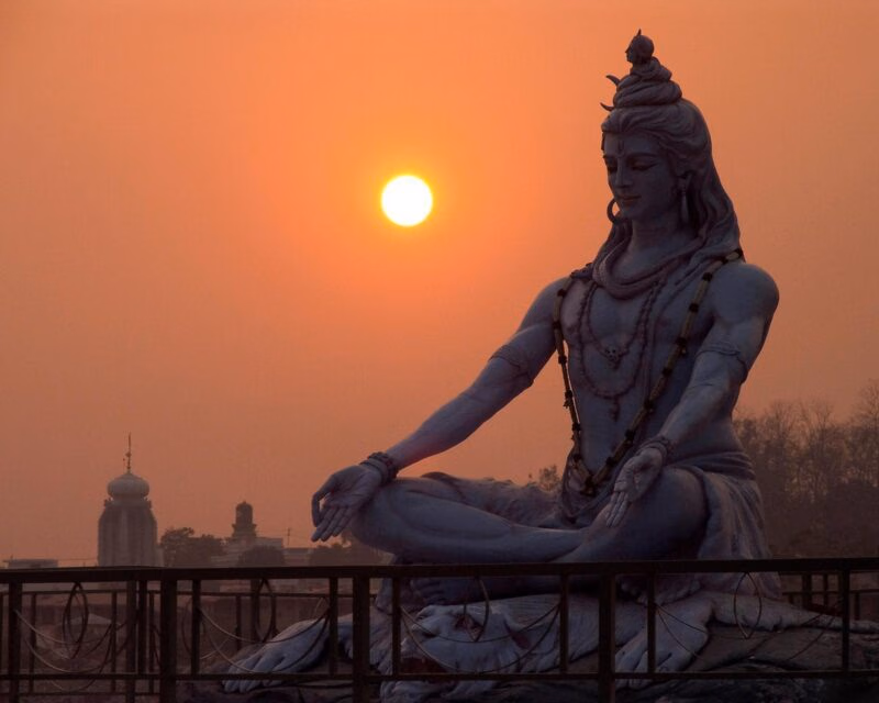 A large statue of Lord Shiva in a seated meditation pose against a glowing orange sunset sky and distant temple.