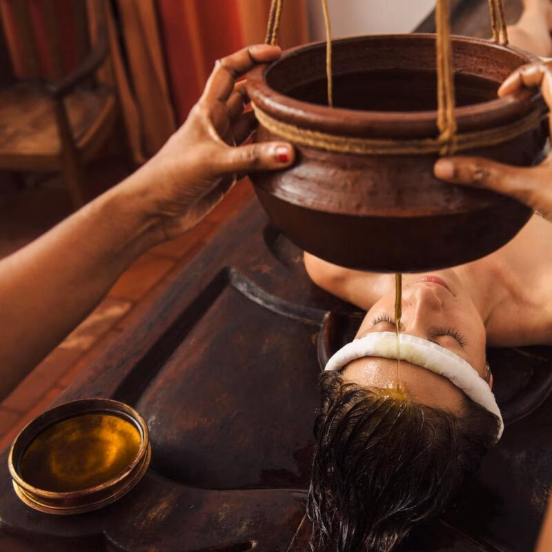 Warm herbal oil pours from a clay pot onto a person's forehead during a traditional Shirodhara Ayurvedic treatment.