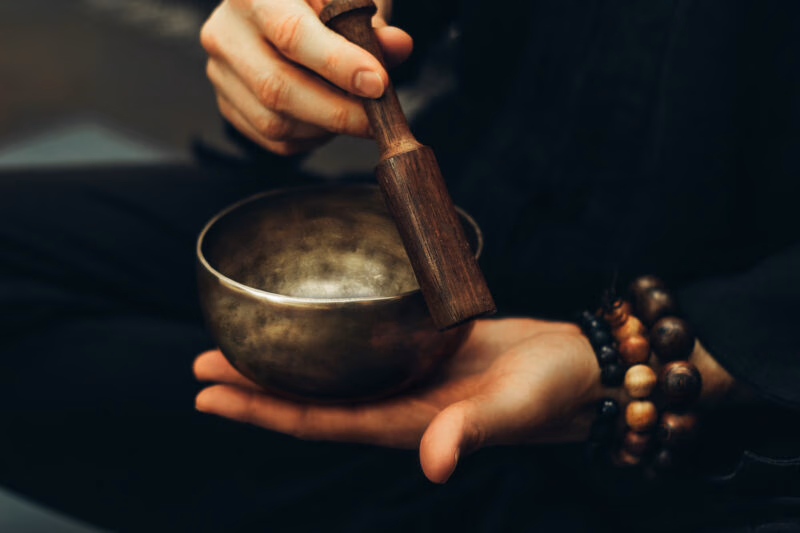 Close-up of a person holding a bronze singing bowl and wooden mallet, wearing beaded bracelets during a sound bath.