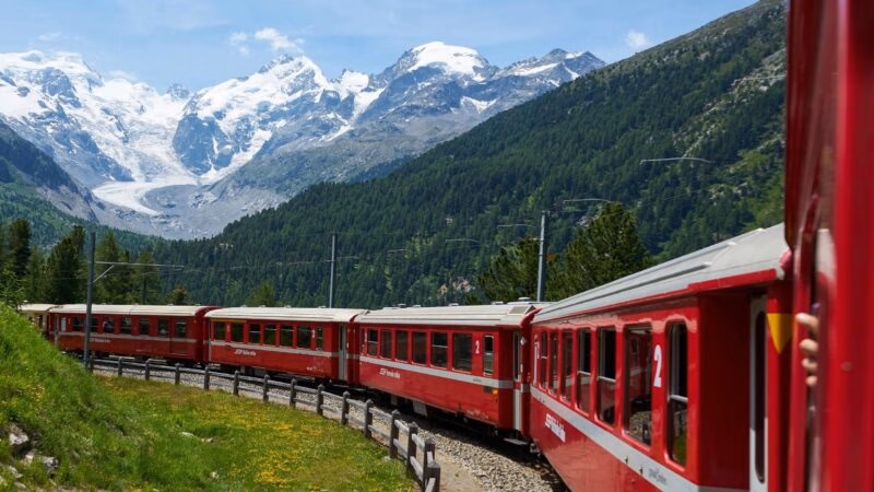 A red Swiss train traveling on a curved track through a mountain valley with snow-covered mountains in the distance.