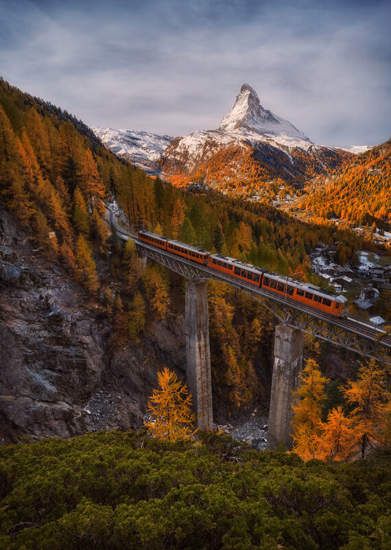 An orange train on a tall bridge surrounded by orange autumn trees with the Matterhorn mountain in the background.