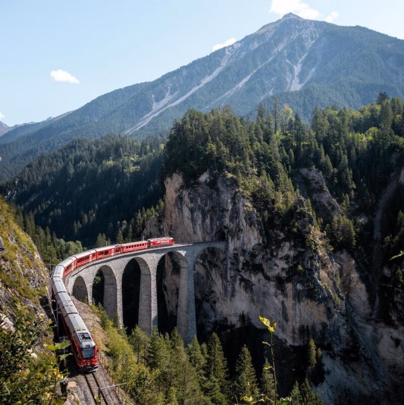 A red train crossing a high arched stone viaduct over a deep forest ravine toward a mountain tunnel.