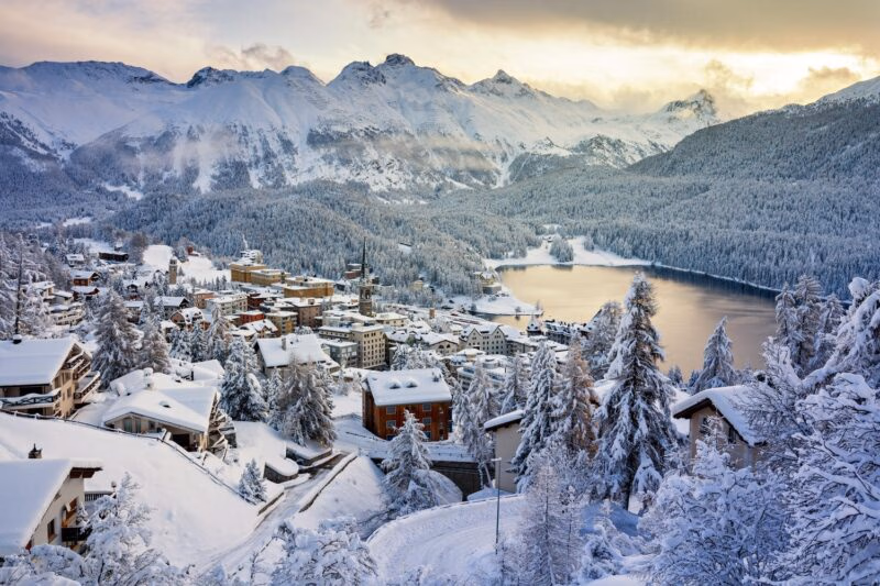 A snowy alpine village and lake in St. Moritz, Switzerland, surrounded by towering mountain peaks at sunset.