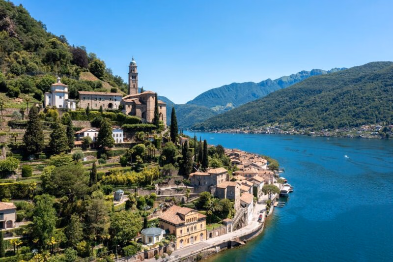 A lakeside village on a lush green hill in Ticino, Switzerland, with blue water and mountains in the distance.