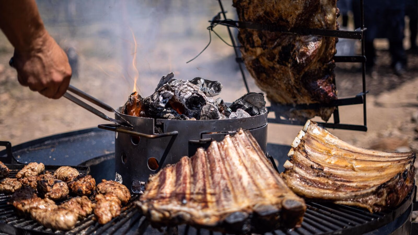Beef ribs and assorted meats grilling over hot coals and flames on a large outdoor metal barbecue.