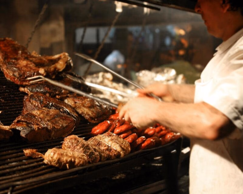 Close-up of a chef grilling steaks and sausages on a large metal barbecue grill with smoke.
