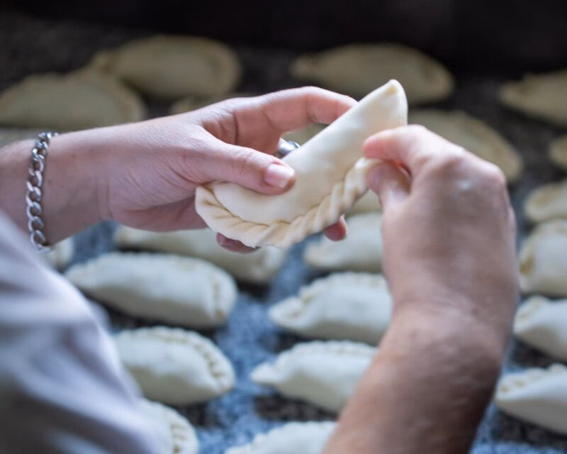 Close-up of hands folding the decorative edge of a raw empanada over a table of prepared pastries.