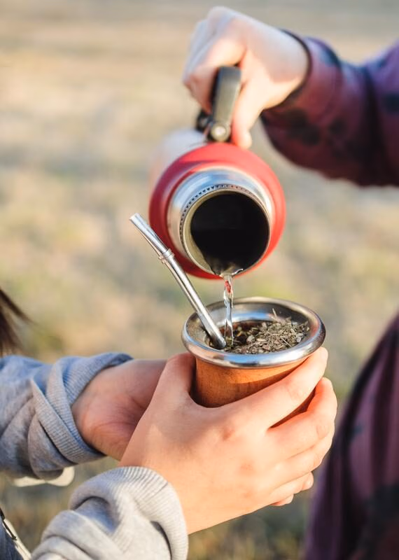 Person pouring water from a thermos into a traditional mate cup with a metal straw held by another person.