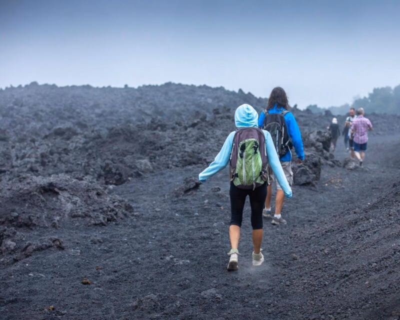 Two hikers with backpacks walk on a trail made of dark volcanic rock toward other people in the distance.