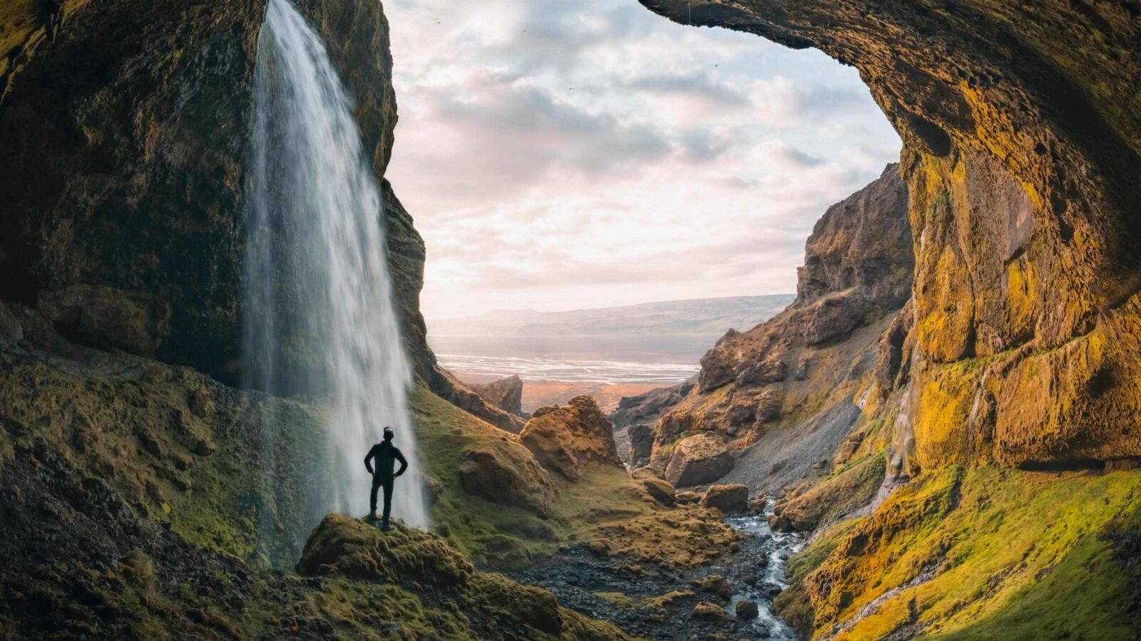 Silhouette of a person standing by a large waterfall flowing from a rocky cave opening overlooking a distant valley.