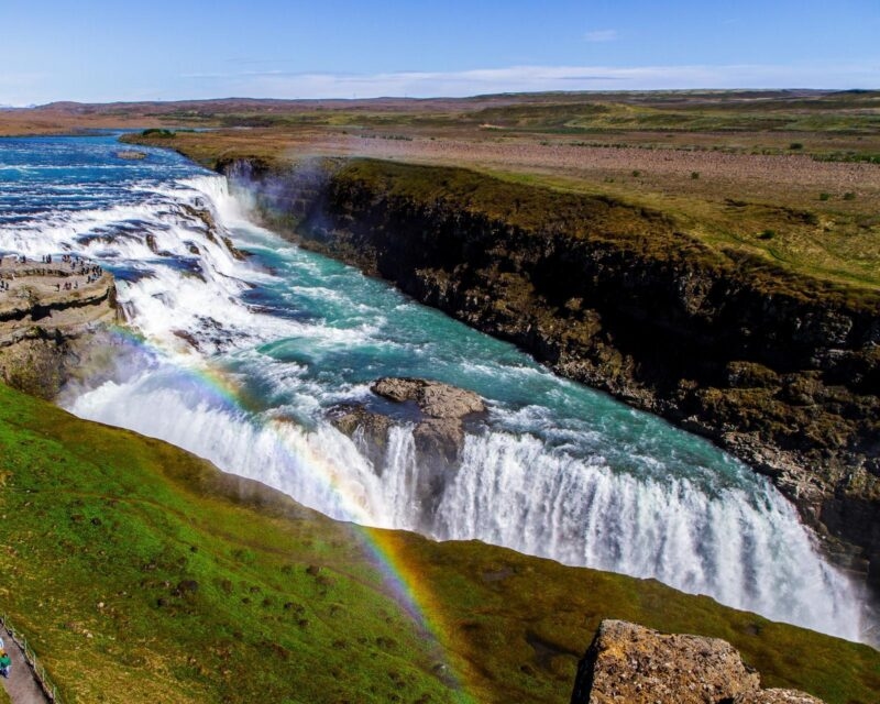 A large multi-tiered waterfall flows into a canyon with a prominent rainbow visible in the mist under a blue sky.