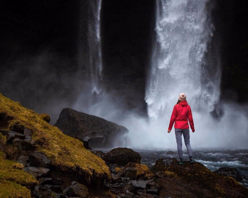 A person in a red jacket stands on dark rocks in front of a tall, misty waterfall flowing into a river.