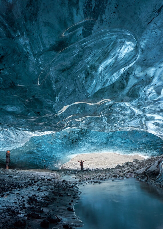 A person stands inside a glowing blue glacial ice cave in Iceland with a small stream running through the center.