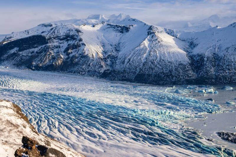 An expansive glacier with blue and white ice ridges sits at the base of snow-covered mountains in Iceland.