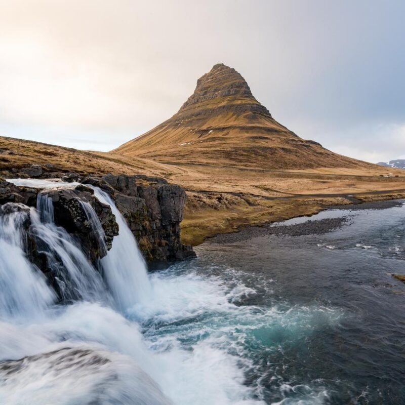 A scenic view of a multi-tiered waterfall in the foreground with a distinct cone-shaped mountain in the background.