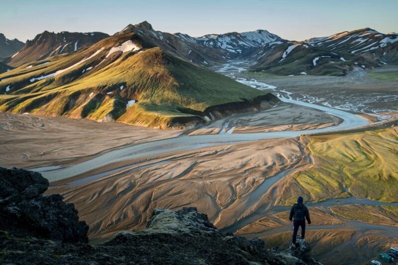 High angle view of a person looking over a wide valley with braided rivers and rolling mountains at golden hour.