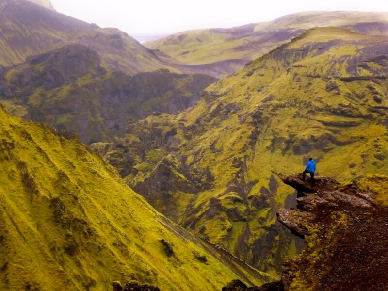 A person in a blue jacket stands on a cliff edge overlooking moss-covered green mountains in Thorsmork, Iceland.