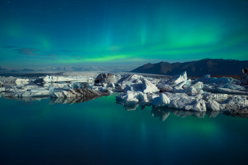 Green northern lights illuminate the night sky above a lagoon with icebergs and distant mountains in Iceland.