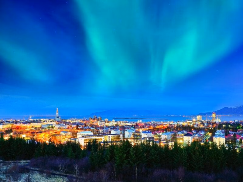 Bright blue and green northern lights stretch across the sky over the illuminated cityscape of Reykjavik, Iceland.