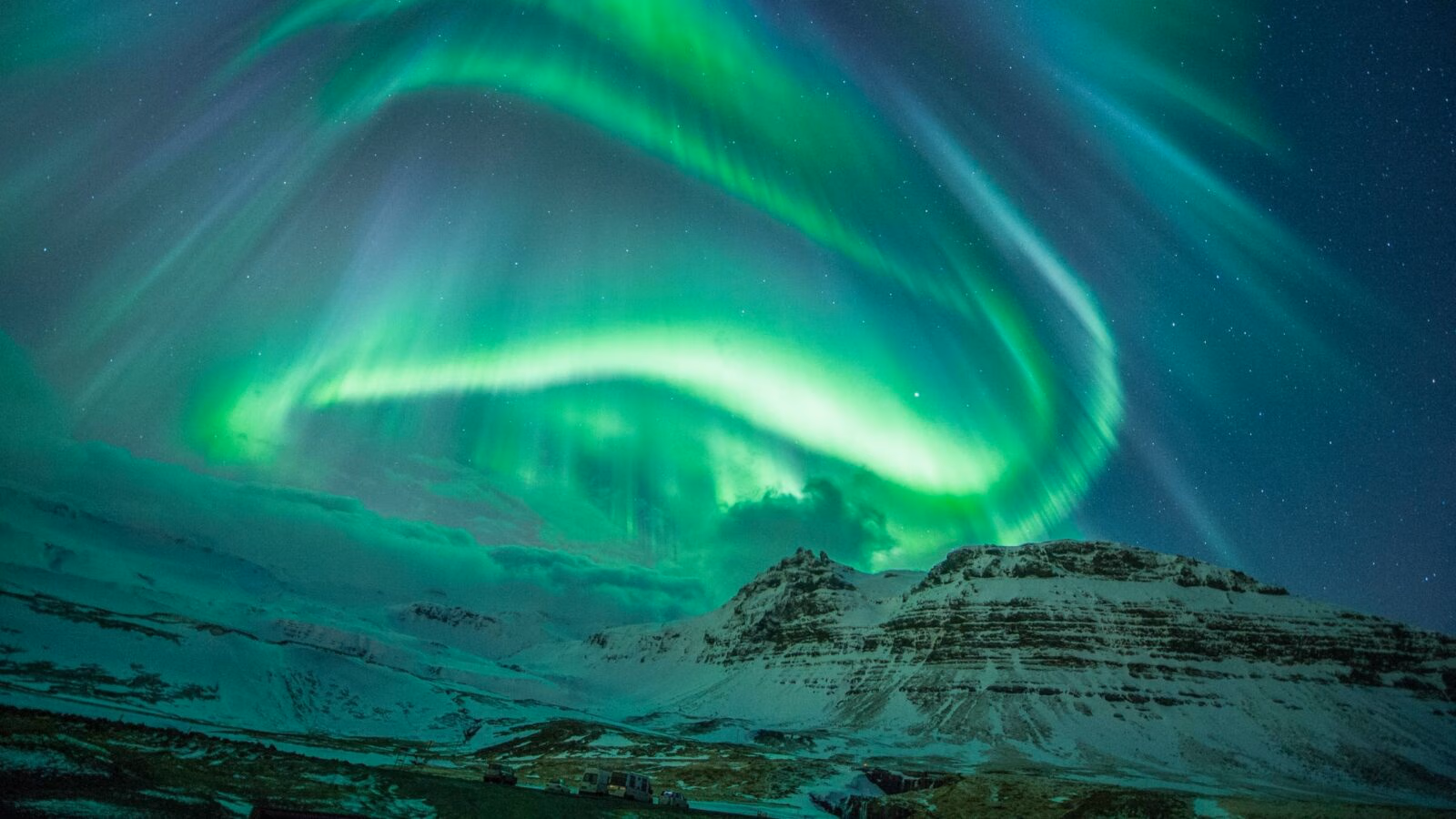 Swirling green aurora borealis lights dance in the night sky over a snowy mountain landscape in Iceland.